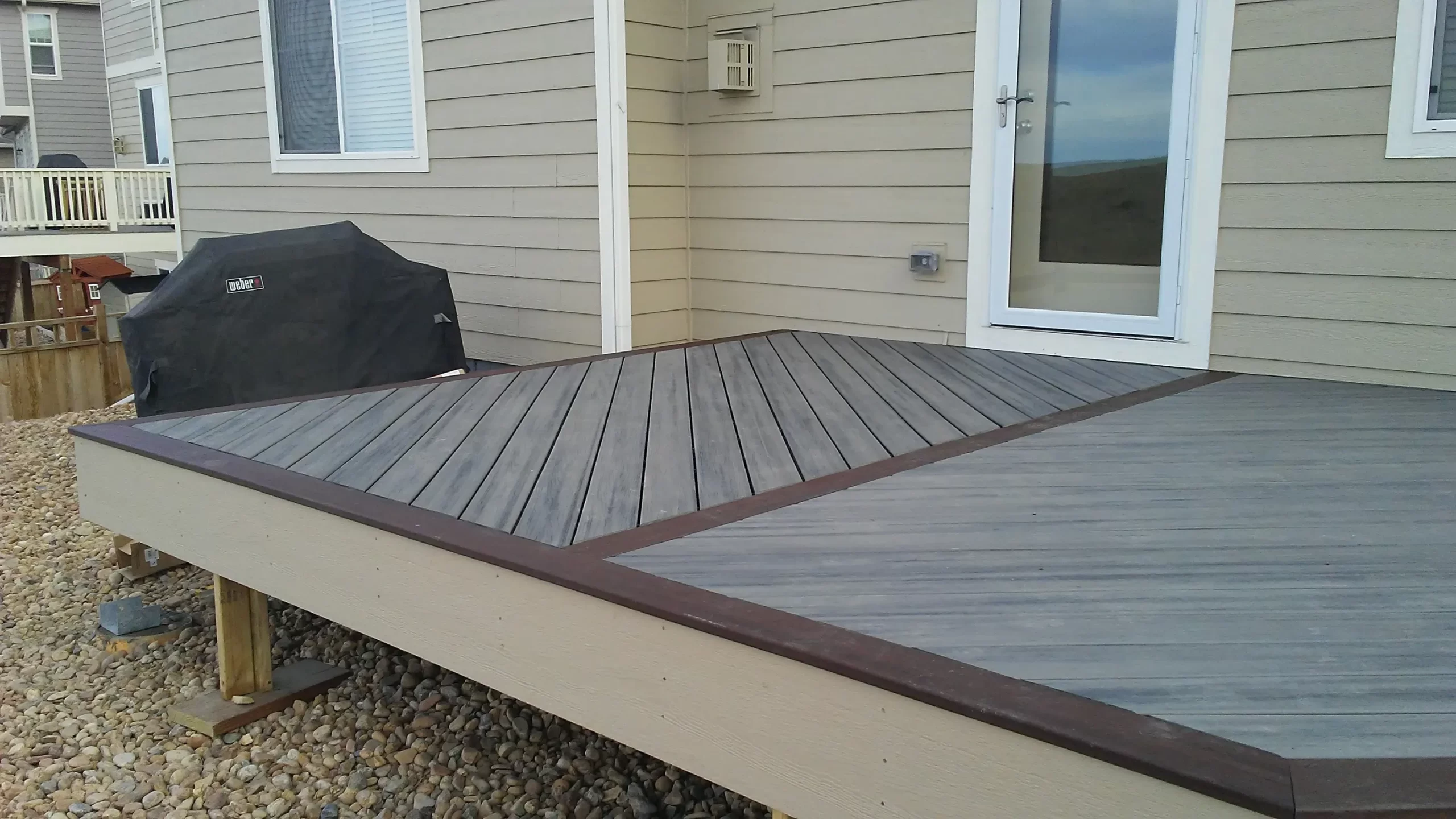 gallery (1) Wooden deck with a unique diagonal pattern, adjacent to a house with a grill covered in black, over pebbled ground.