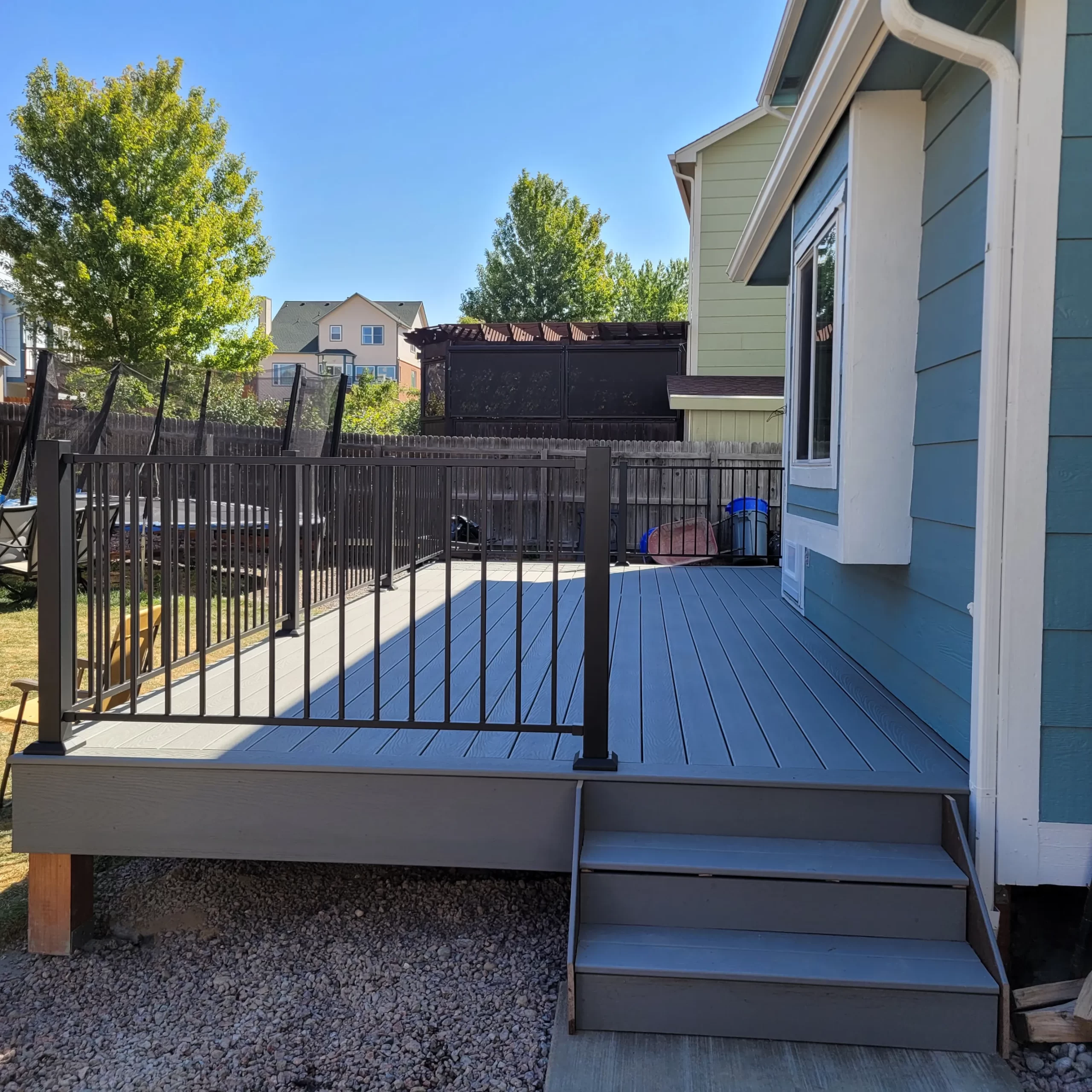 gallery (12) A wooden backyard deck with black railings and stairs, connected to a blue house on a sunny day.