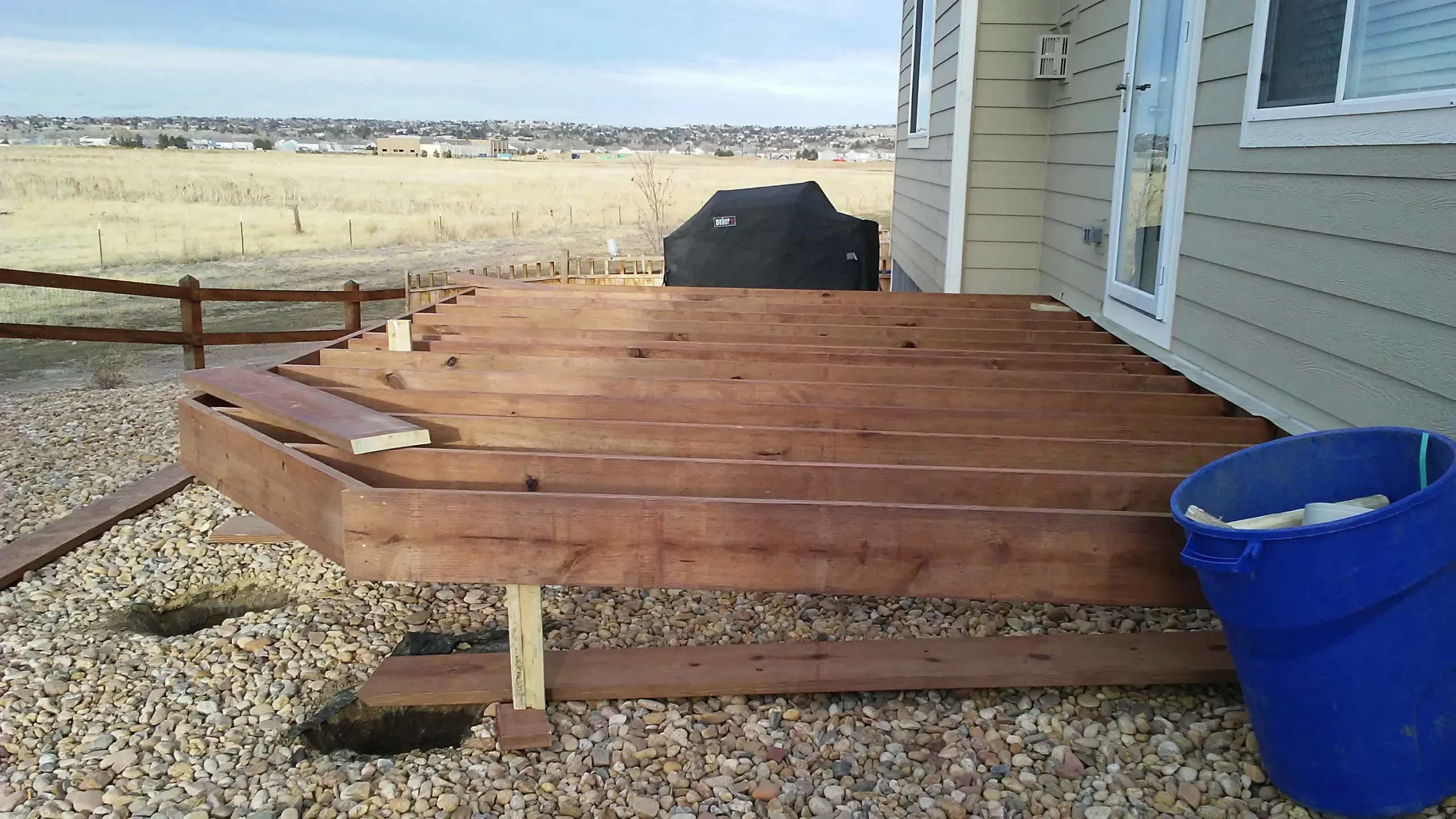 gallery (17) A wooden deck under construction in a backyard, with planks and tools visible, adjacent to a house.