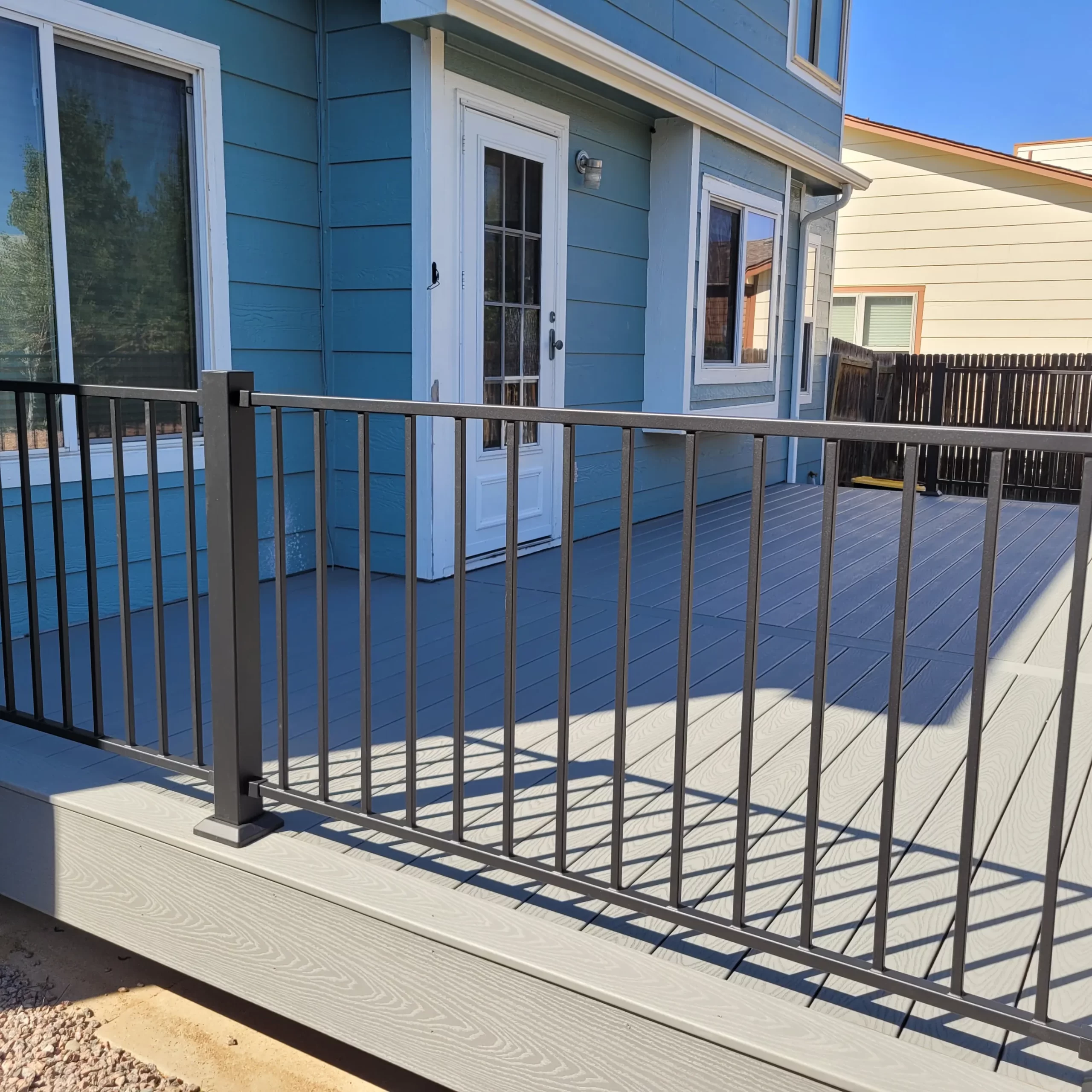 gallery (20) Blue house with a white door and a modern deck featuring black metal railing, set against a clear blue sky.