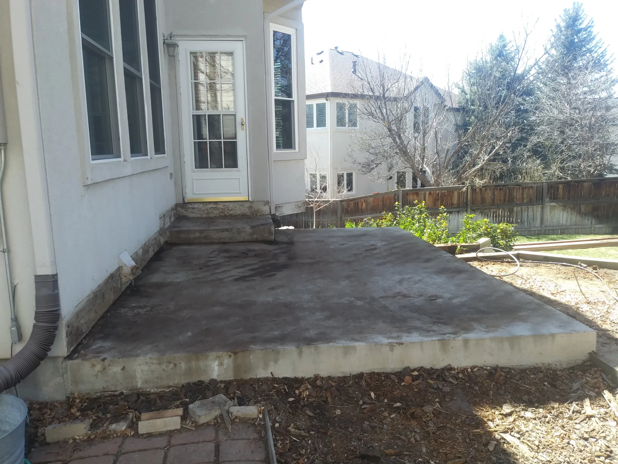 gallery (22) Concrete patio with steps leading to a white door, surrounded by a wooden fence and trees in the background.