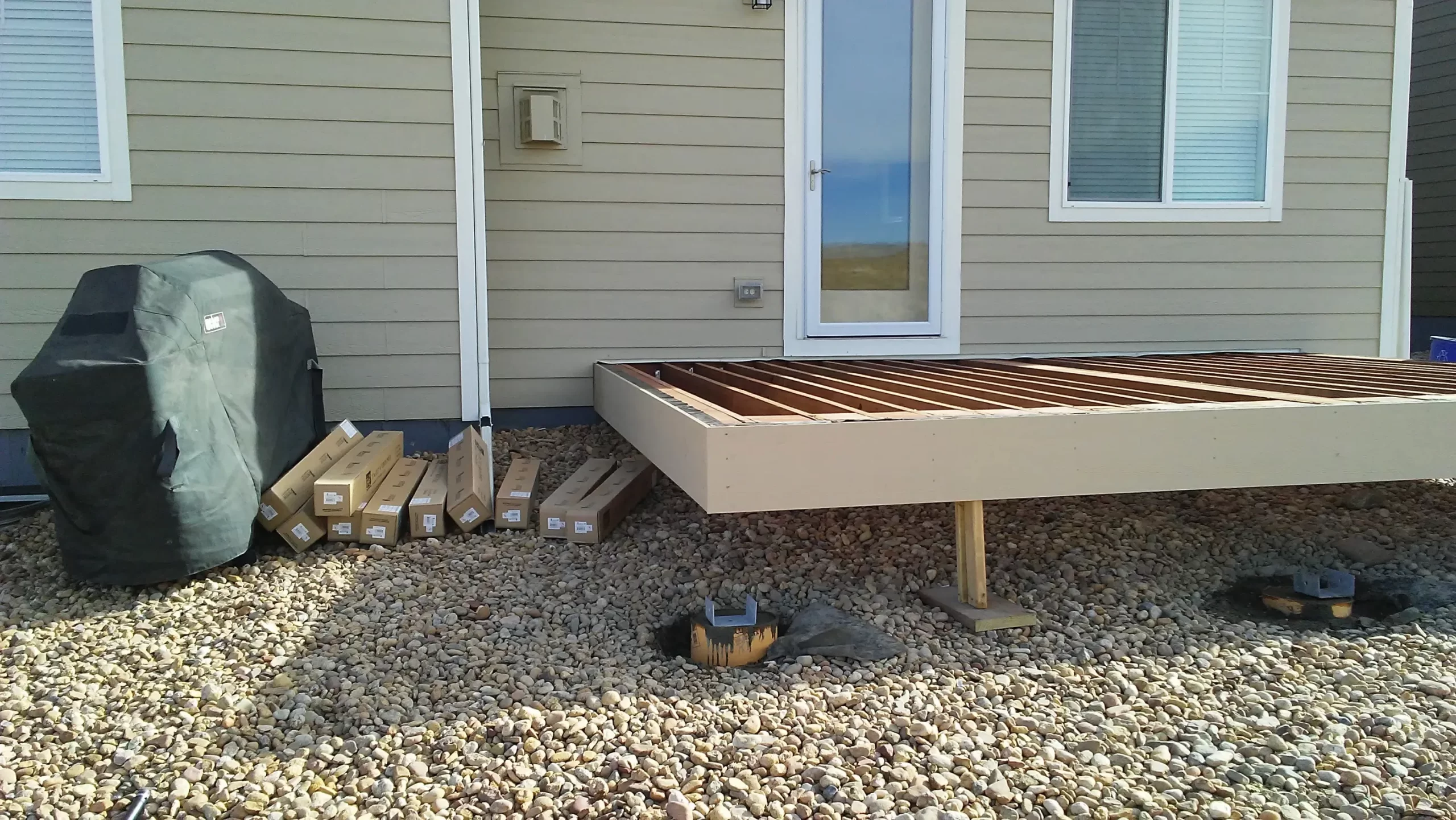 gallery (23) Partially constructed deck with wooden beams, surrounded by gravel, near a beige house with a covered grill and boxes.
