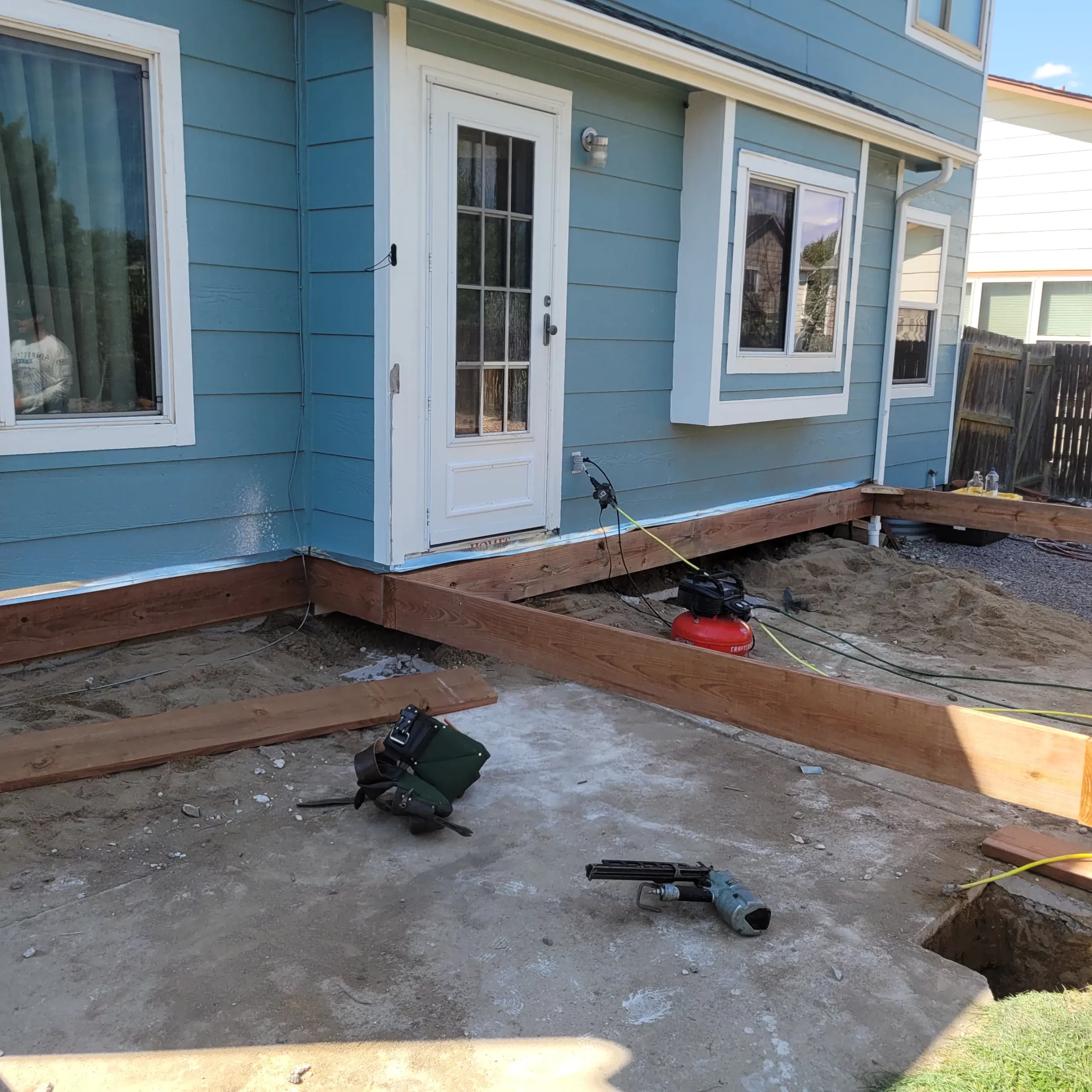 gallery (26) Backyard construction site with wooden beams on concrete, blue house exterior, door, and a compressor.