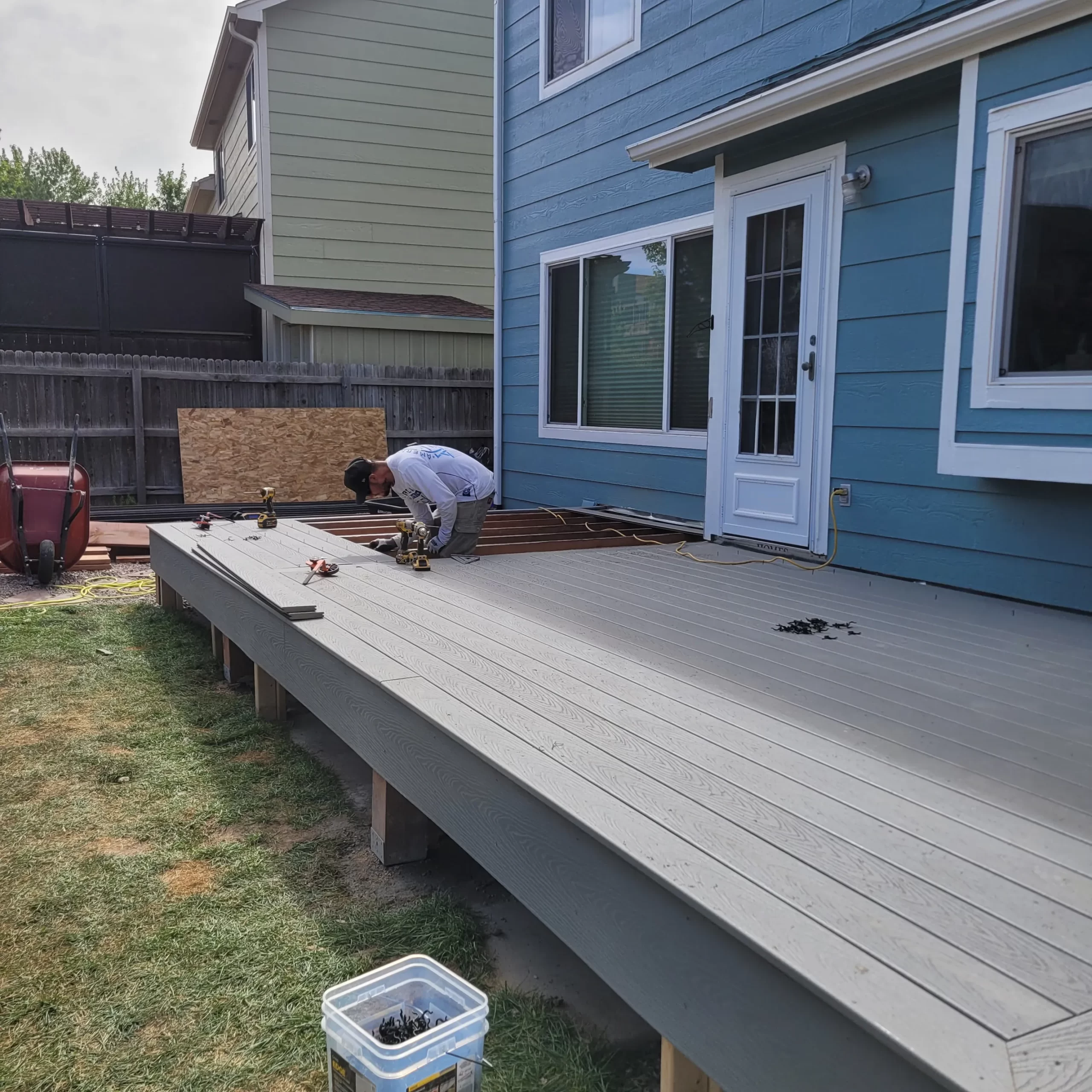 gallery (27) Person working on building a raised deck attached to a blue house in a backyard.