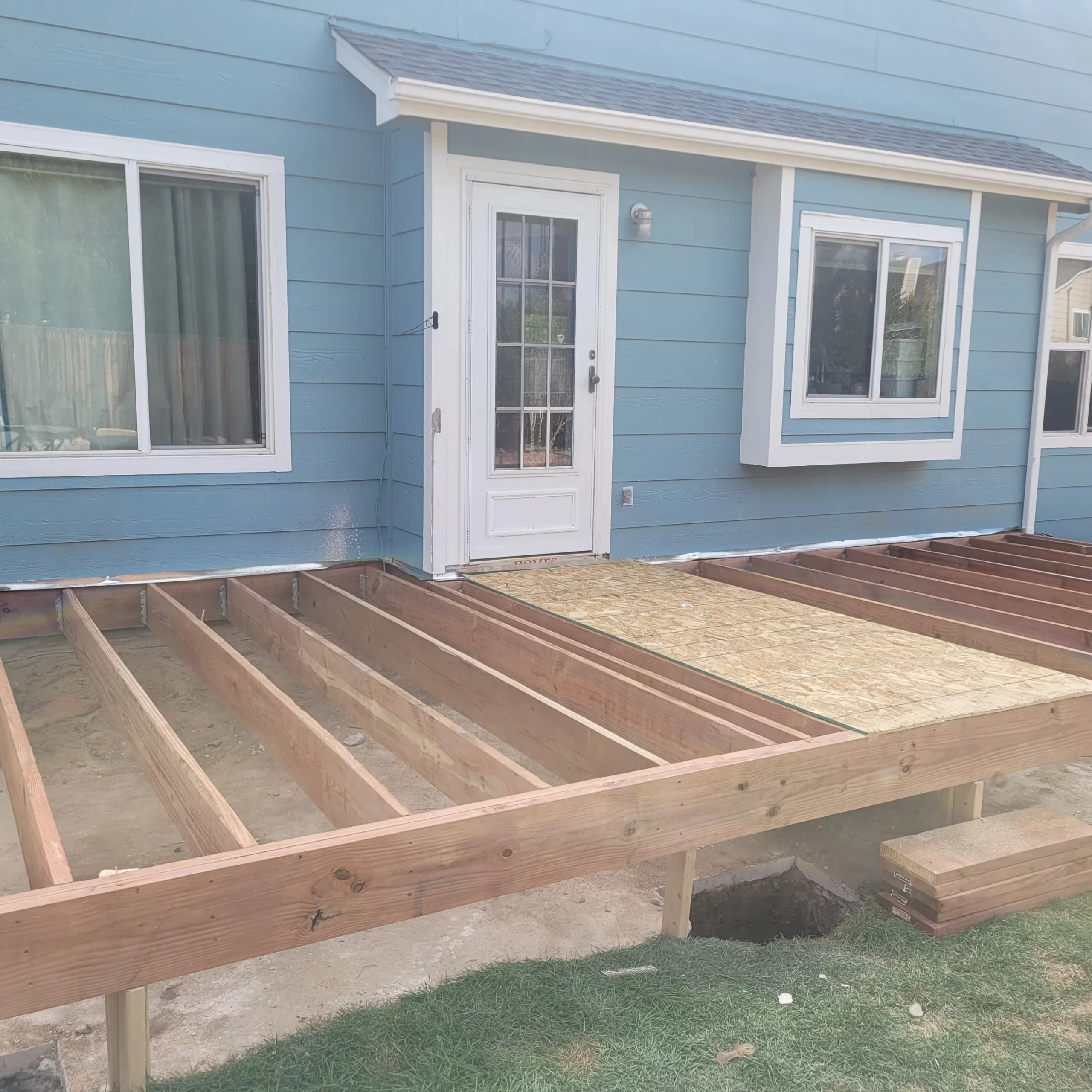 gallery (3) Wooden deck frame under construction against a blue house with a white door and two windows.