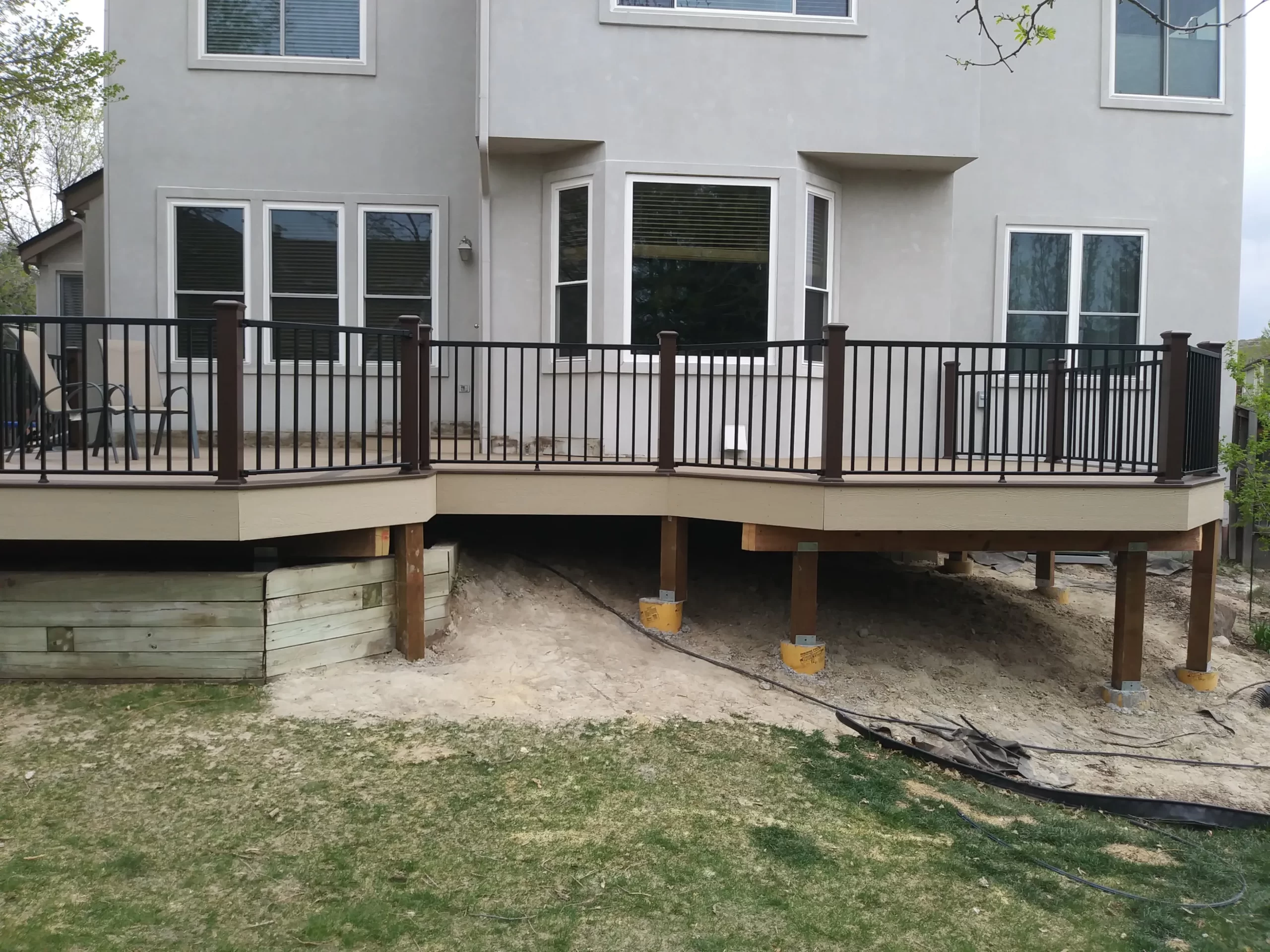 gallery (4) A beige house with a raised deck featuring dark railing, supported by wooden posts, over a sandy ground.