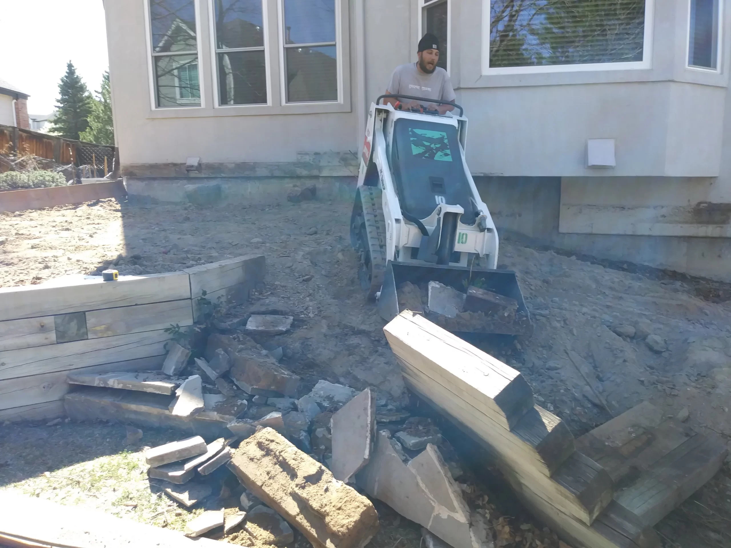 gallery (5) Man operating a small skid-steer loader, moving debris in a yard near a house.