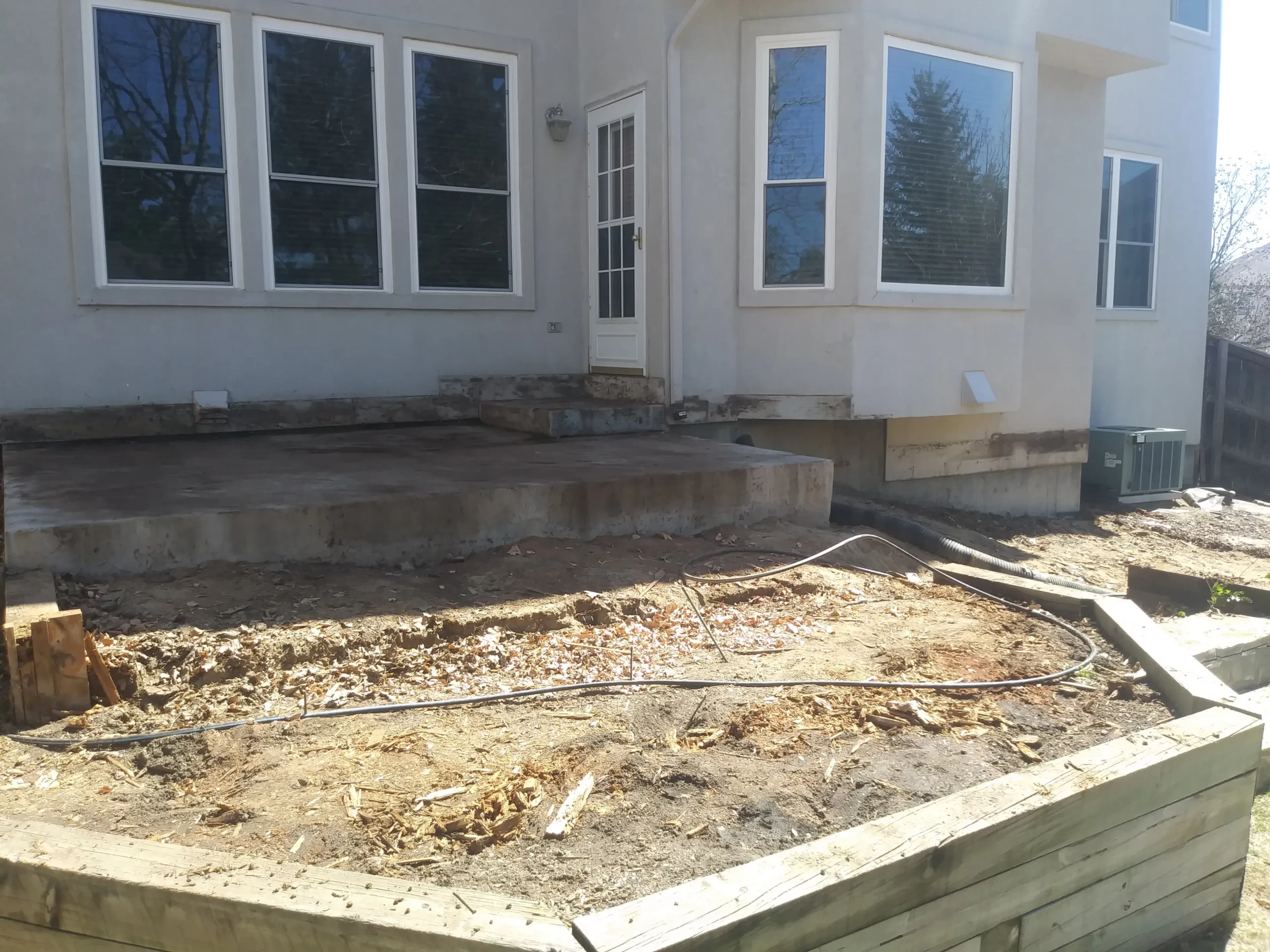 gallery (6) Backyard with a raised patio, dirt area, and retaining wall, in front of a house with several large windows.