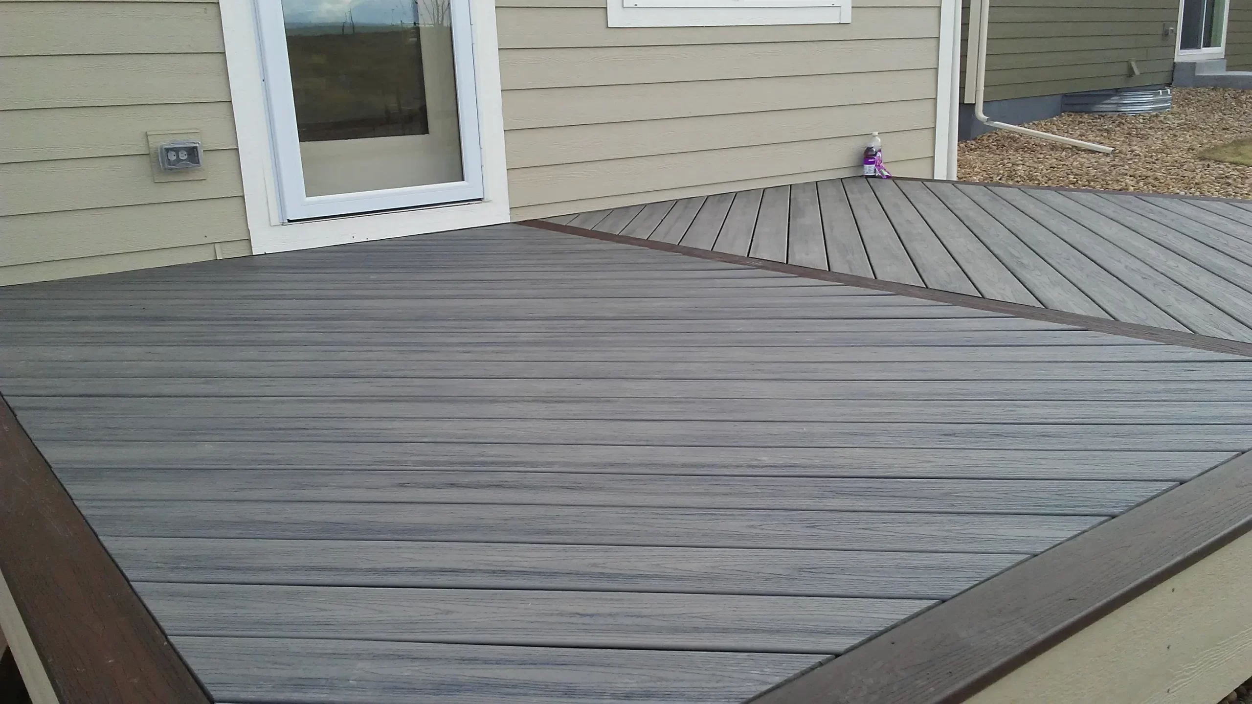 gallery (7) Wooden deck with diagonal planks in front of a beige house. A glass door and small purple object are visible.