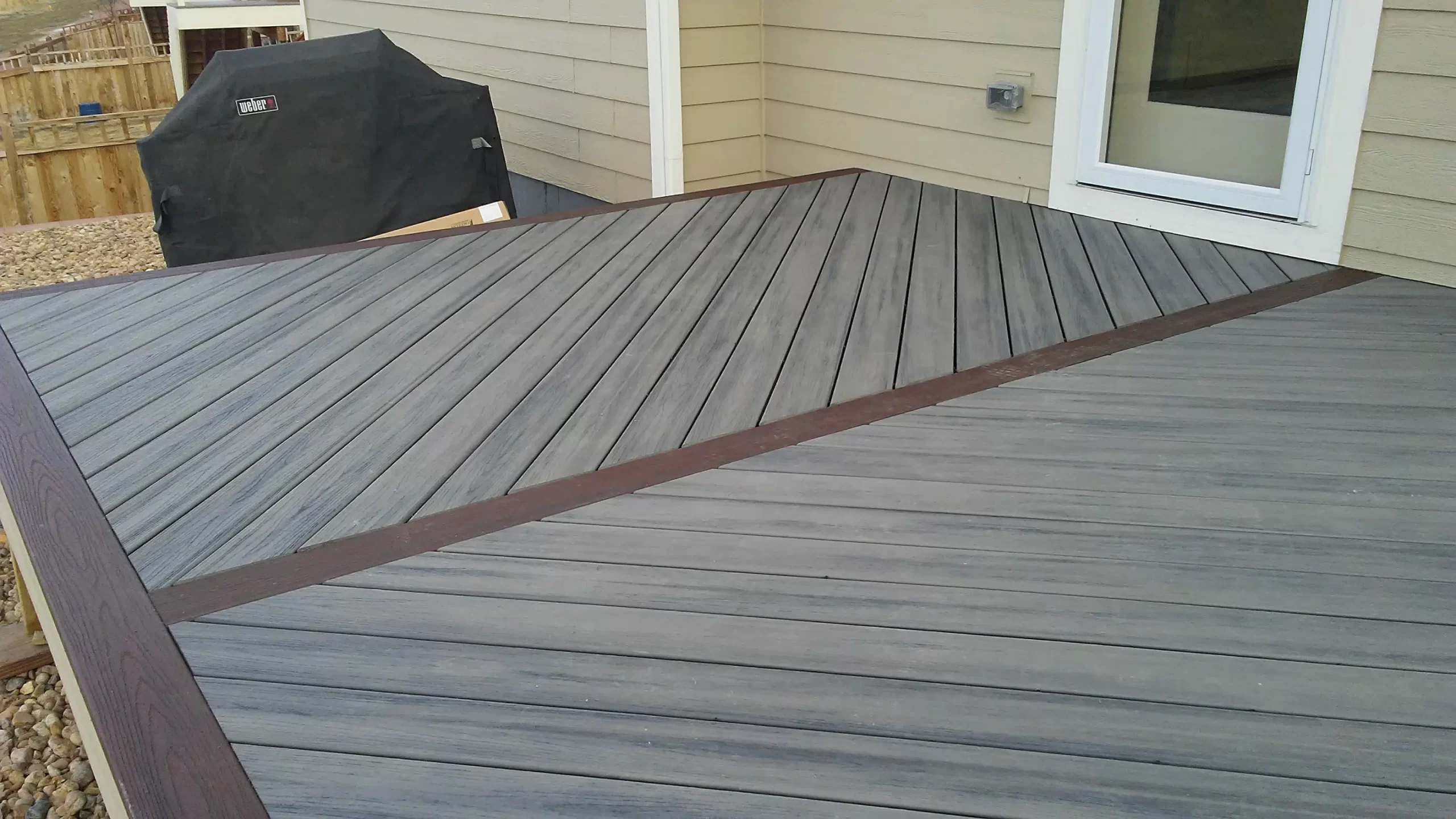 gallery (8) Wooden deck with diagonal planks near a beige house and a covered grill, leading to a glass door.
