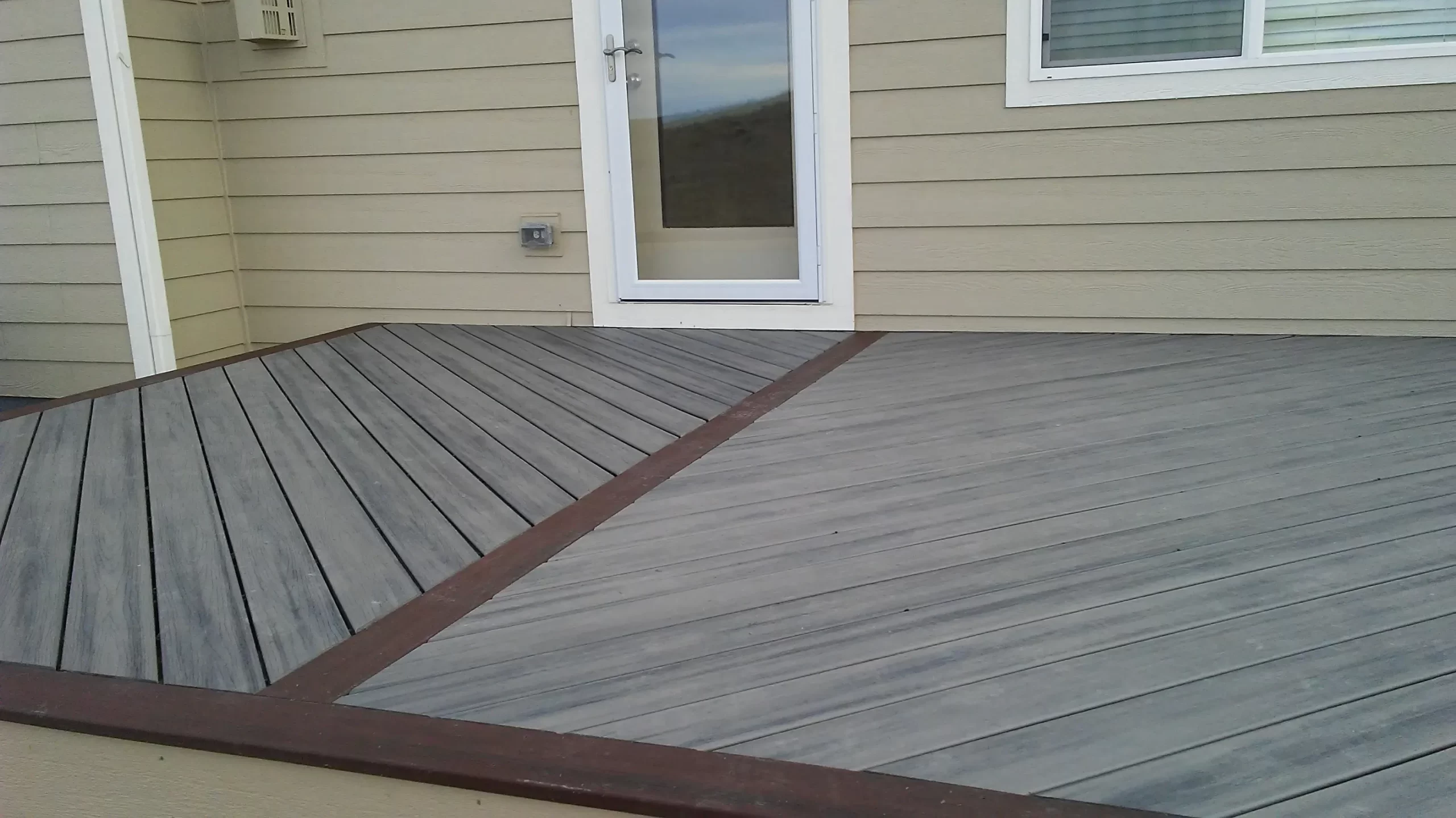 gallery (9) Wooden deck with angled patterns, adjacent to a beige paneled exterior wall and glass door.