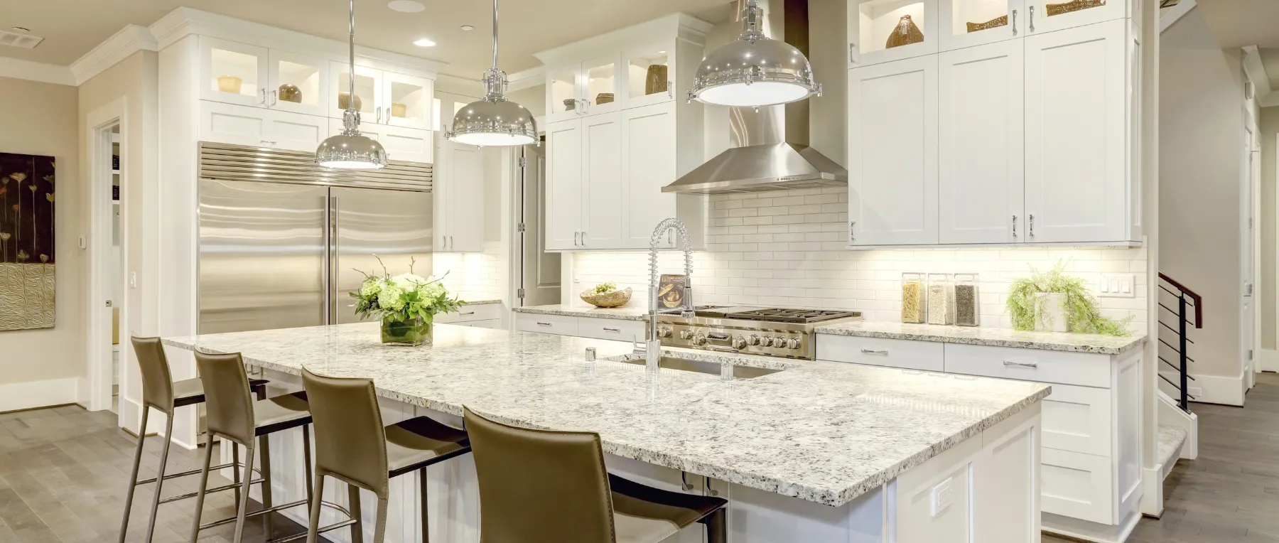 Modern kitchen with a large marble island, pendant lights, stainless steel appliances, and four brown chairs.
