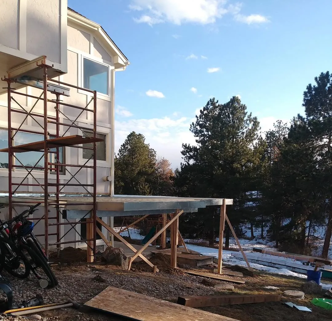 about-1 House under construction with scaffolding and a deck frame, set against a backdrop of trees and a clear sky.