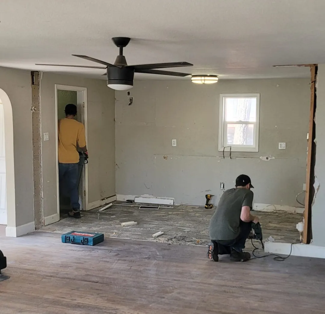 about-2 Two people working on home renovations, with exposed walls and floors, under a ceiling fan.