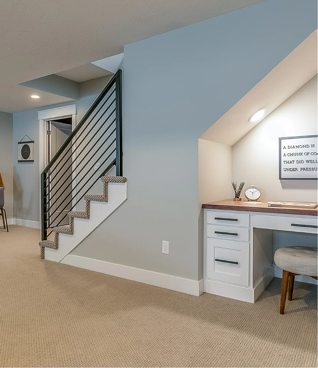 basement-finishing-2 Home office nook under stairs with desk, drawers, a chair, and wall art. Carpeted floor and a modern railing staircase.