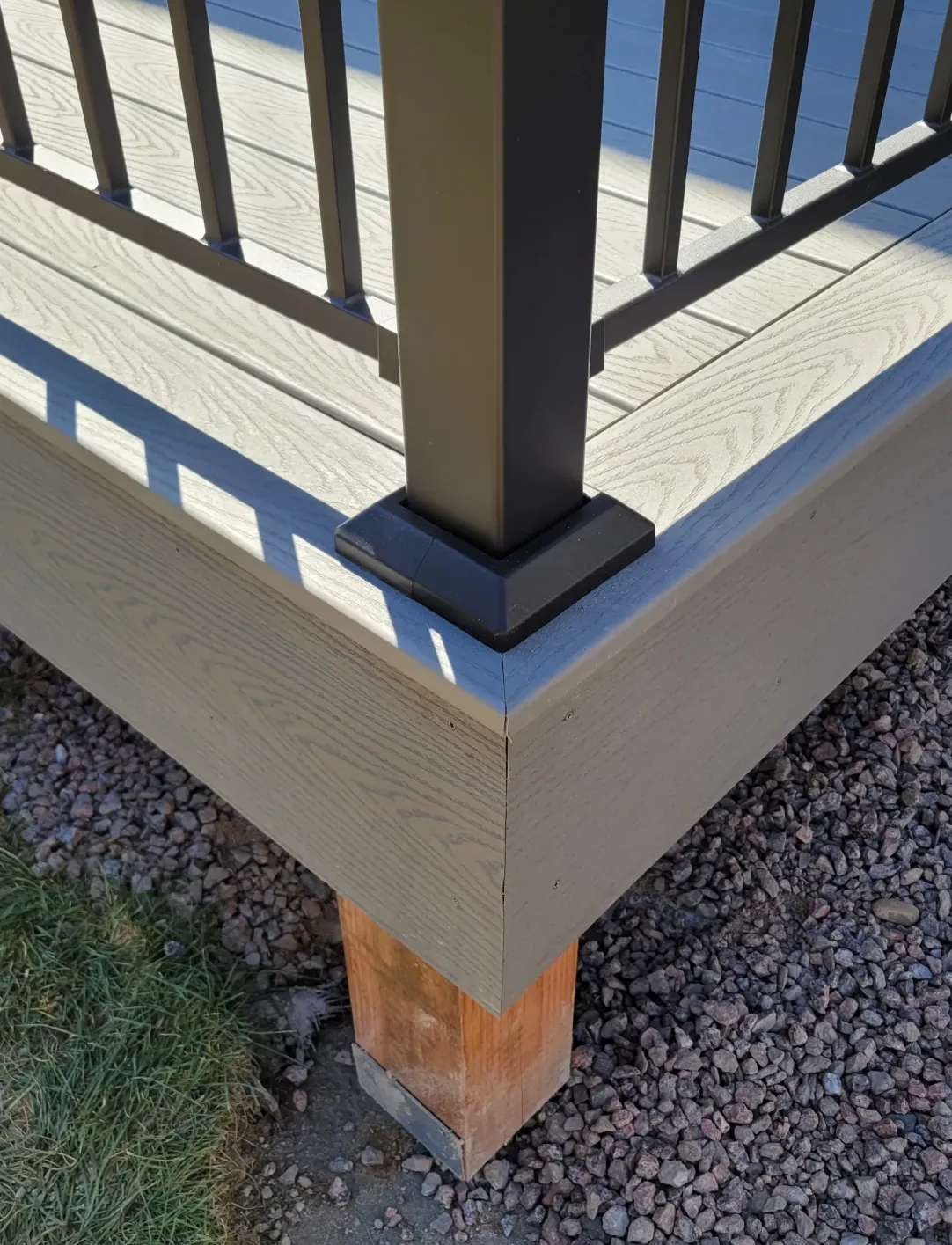 deck-repair-1 Corner of a gray composite deck with black metal railing on a wooden post foundation, surrounded by gravel.