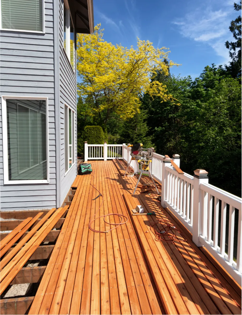 deck-repair-2 A person works on a wooden deck repair next to a gray house, surrounded by trees under a clear blue sky.