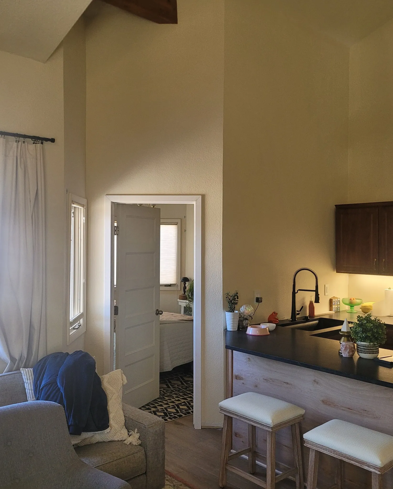 home-remodeling-1 Cozy living area with sofa, open door to bedroom, and kitchen counter with bar stools. Warm lighting throughout.