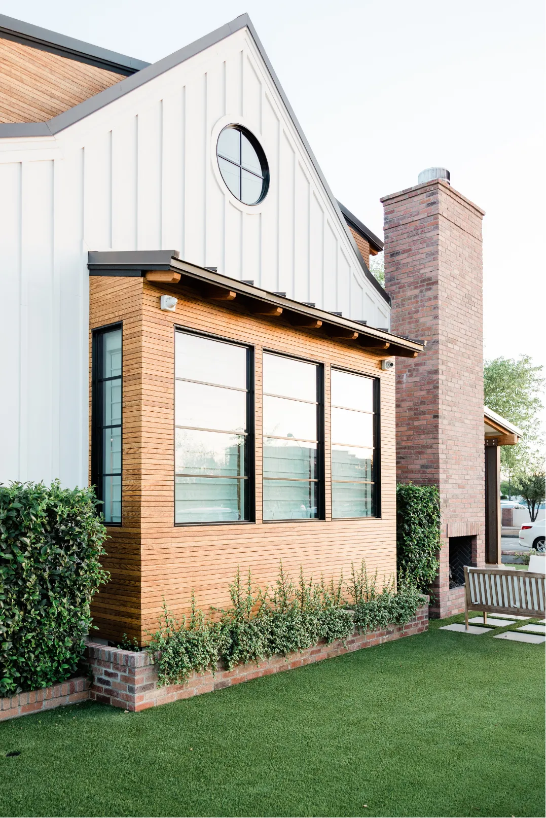 home-remodeling-2 Modern house exterior with white siding, circular window, and brick chimney, beside manicured lawn and bench.
