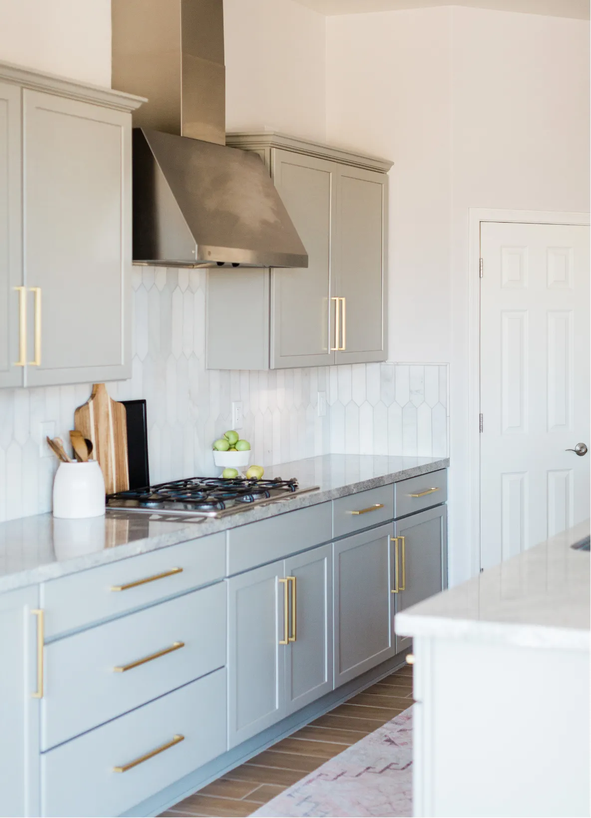 kitchen-remodeling-3 Modern kitchen with gray cabinets, gold handles, and white backsplash. Features stove, vent hood, and countertop decor.