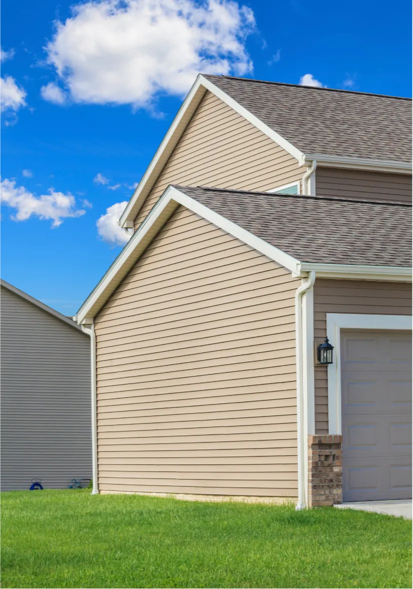 Close-up of a beige house with a gable roof, white trim, grassy lawn, and a bright blue sky with clouds.