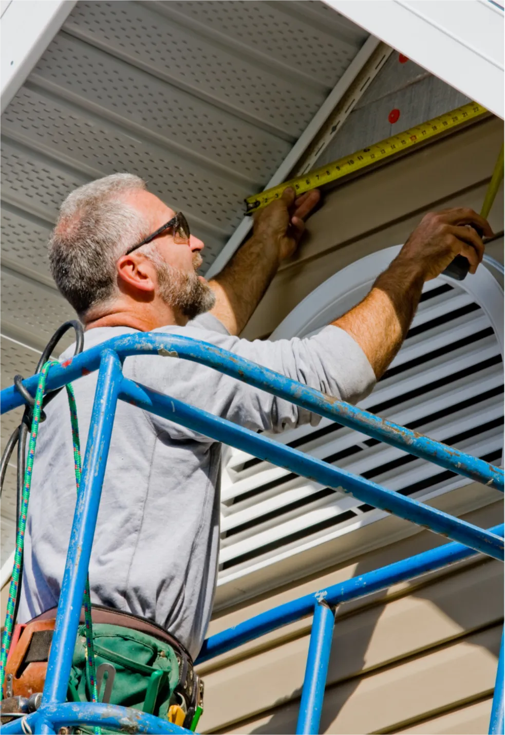siding-repairs-2 Man on blue scaffold measures house exterior with a tape measure, wearing a tool belt and sunglasses.