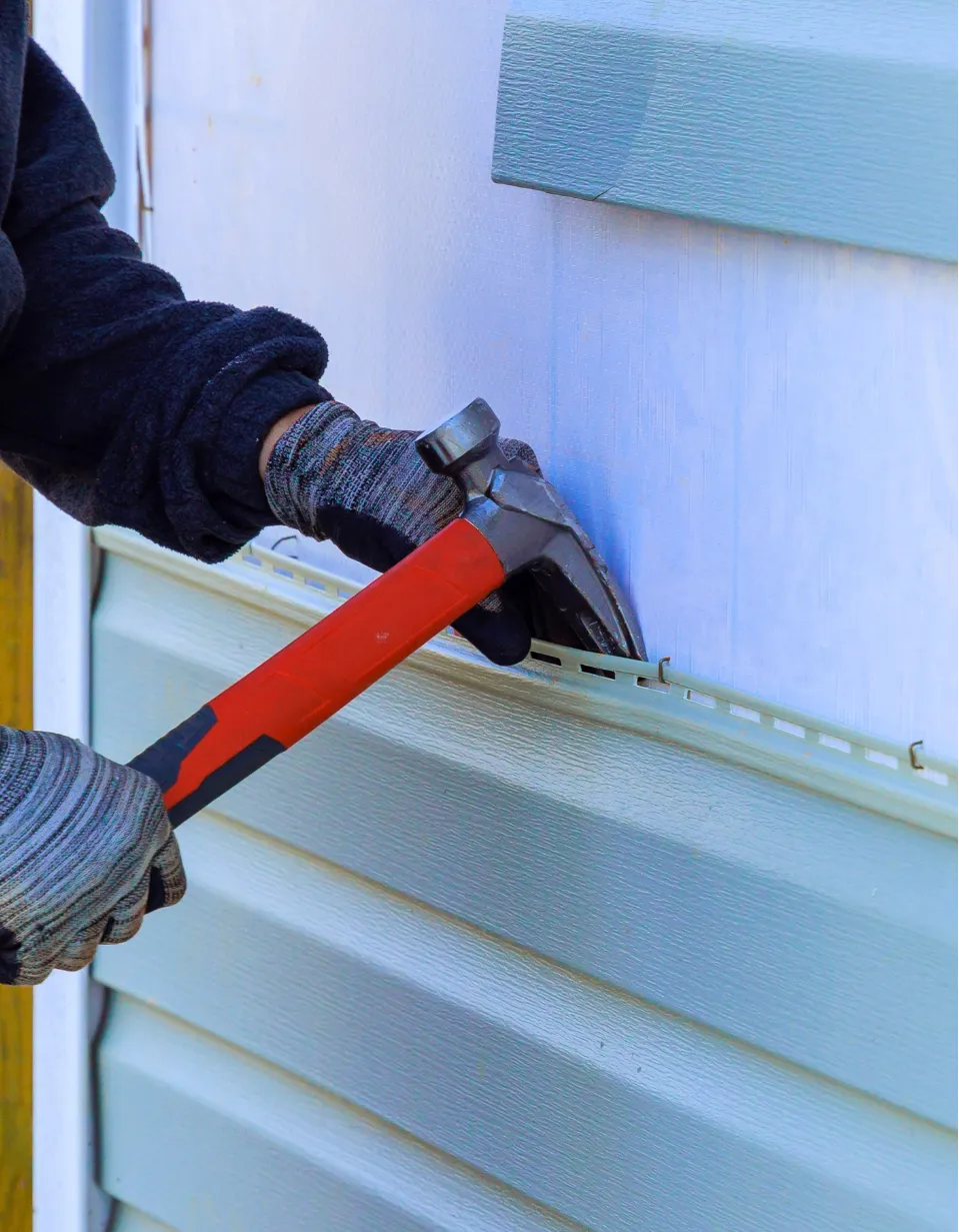 siding-replace-2 Person wearing gloves using a hammer to install blue Siding Installation or siding replacement on a wall.