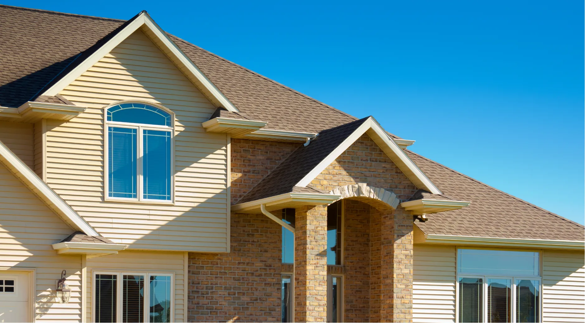 siding-replace-h Modern house with a brown shingle roof, arched windows, and beige siding under a clear blue sky.
