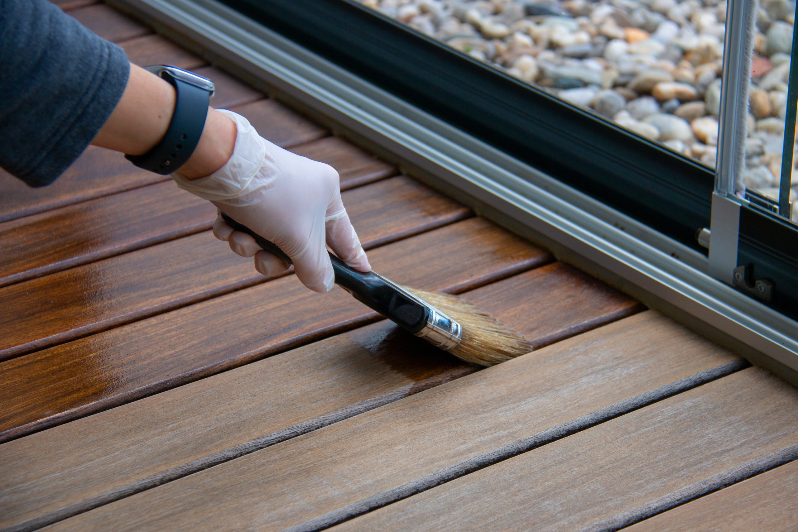 Deck staining, worker applying deck oil on decking boards with paint brush, hardwood terrace renovation Person with glove and watch applying varnish to wooden deck with brush, pebbles visible in background.