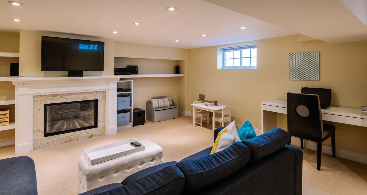 A cozy basement living room with a TV, fireplace, desk, and blue sofa under recessed lighting.
